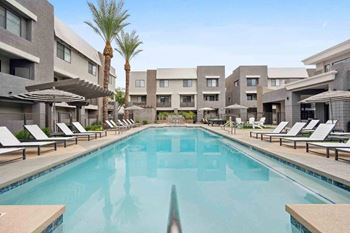 A large swimming pool surrounded by lounge chairs and umbrellas in front of apartment buildings.